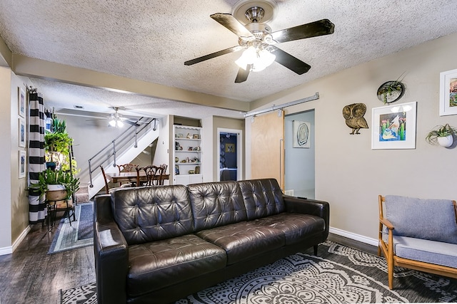 living room with built in features, ceiling fan, a barn door, dark wood-type flooring, and a textured ceiling