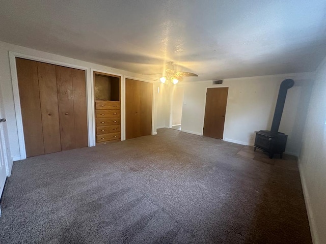 unfurnished bedroom with two closets, carpet floors, a wood stove, and ceiling fan