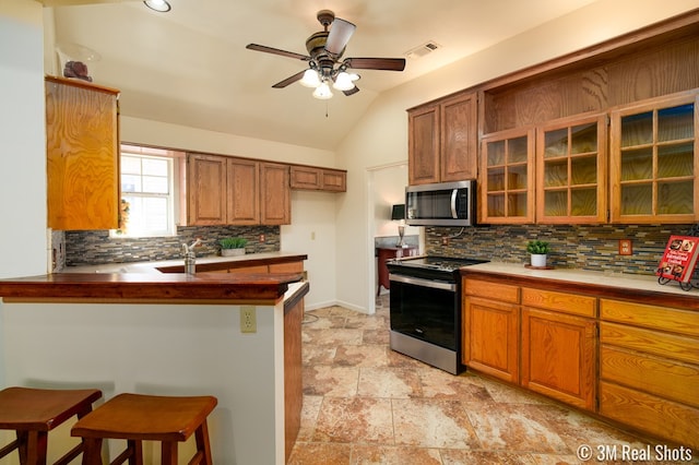 kitchen featuring glass insert cabinets, vaulted ceiling, appliances with stainless steel finishes, a kitchen breakfast bar, and brown cabinetry