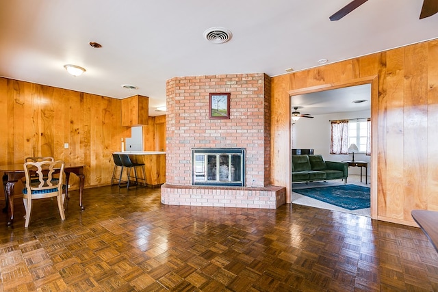 unfurnished living room featuring a ceiling fan, a brick fireplace, visible vents, and wooden walls