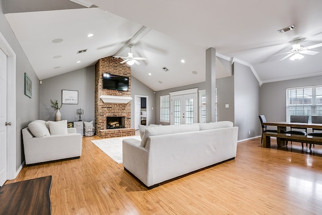 living room featuring ceiling fan, vaulted ceiling with beams, ornamental molding, a brick fireplace, and light wood-type flooring