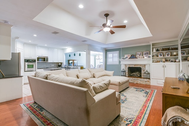 living room featuring a fireplace, ornamental molding, ceiling fan, a tray ceiling, and light hardwood / wood-style flooring