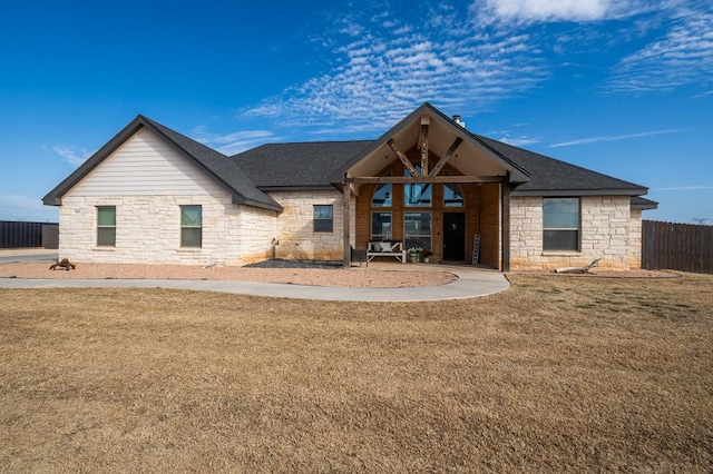 back of house featuring stone siding, a shingled roof, fence, and a lawn