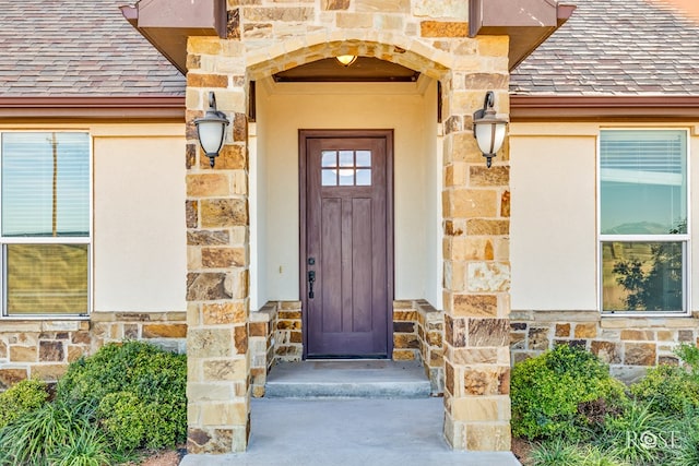 view of exterior entry with stone siding, stucco siding, and a shingled roof