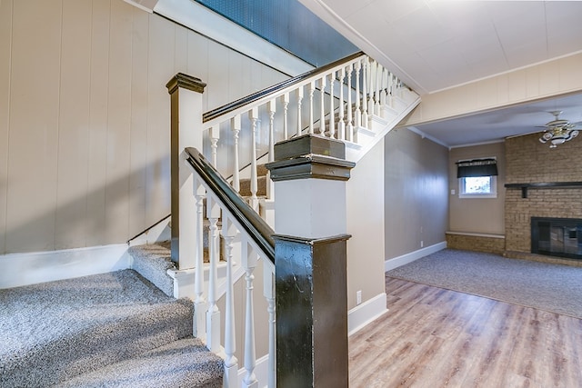 stairway with crown molding, a brick fireplace, ceiling fan, wood finished floors, and baseboards