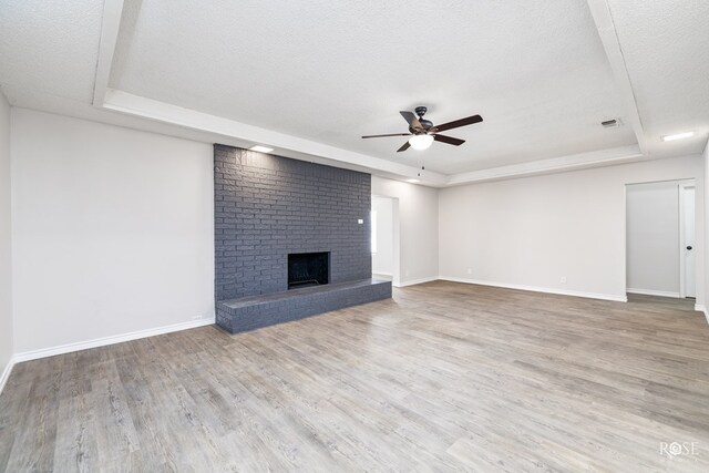 unfurnished living room with a brick fireplace, a textured ceiling, a raised ceiling, ceiling fan, and light hardwood / wood-style floors