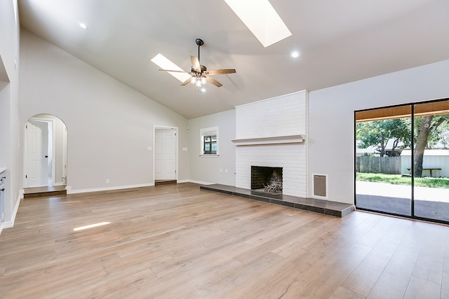 unfurnished living room featuring high vaulted ceiling, a skylight, ceiling fan, a brick fireplace, and light hardwood / wood-style flooring