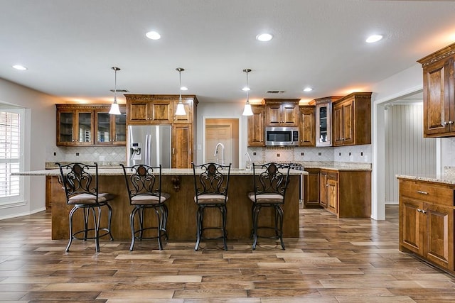 kitchen with hanging light fixtures, appliances with stainless steel finishes, an island with sink, light stone countertops, and hardwood / wood-style floors