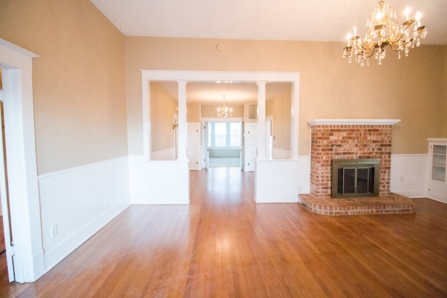 unfurnished living room featuring a notable chandelier, a wainscoted wall, a brick fireplace, ornate columns, and wood finished floors