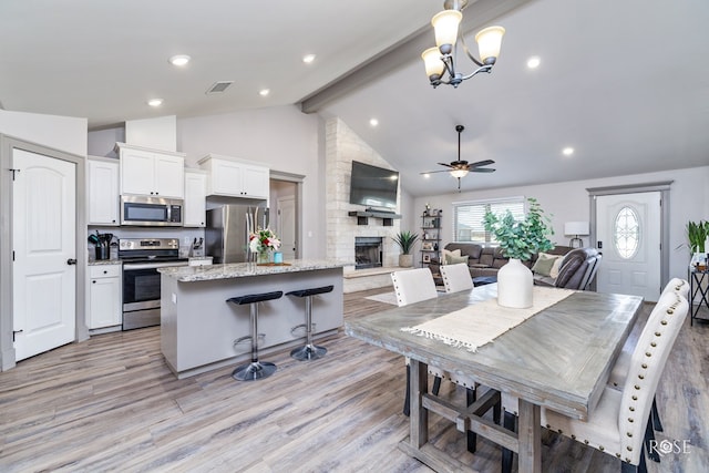 dining space featuring high vaulted ceiling, beam ceiling, light wood-style flooring, a stone fireplace, and ceiling fan with notable chandelier