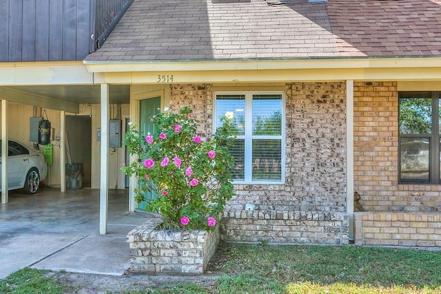view of exterior entry featuring a carport, a shingled roof, electric panel, and brick siding