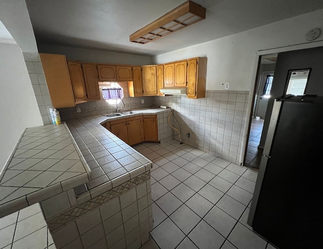 kitchen featuring tile counters, freestanding refrigerator, a sink, a peninsula, and under cabinet range hood