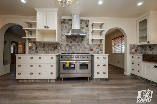 kitchen with range with two ovens, wall chimney range hood, white cabinets, and backsplash