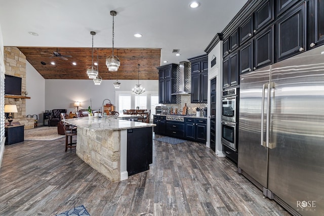 kitchen with wall chimney range hood, appliances with stainless steel finishes, an island with sink, decorative light fixtures, and wooden ceiling