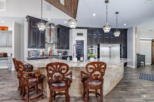 kitchen featuring a breakfast bar area, a large island with sink, hanging light fixtures, stainless steel appliances, and wall chimney exhaust hood