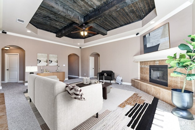 carpeted living room featuring a towering ceiling, a tiled fireplace, a raised ceiling, crown molding, and beam ceiling