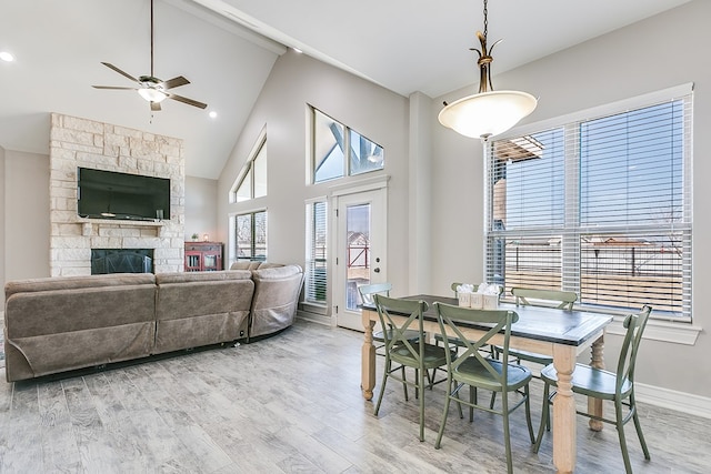 dining area featuring baseboards, light wood-style flooring, a fireplace, high vaulted ceiling, and a ceiling fan