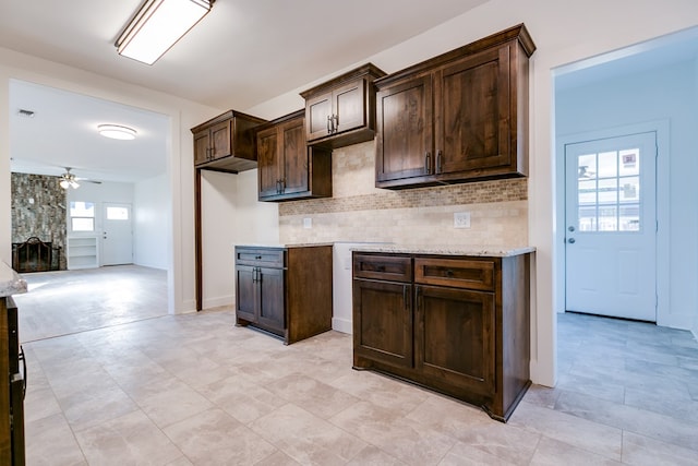 kitchen featuring ceiling fan, dark brown cabinetry, a fireplace, light stone countertops, and decorative backsplash