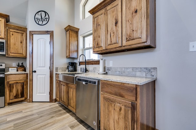 kitchen with appliances with stainless steel finishes, brown cabinetry, a sink, light stone countertops, and light wood-type flooring