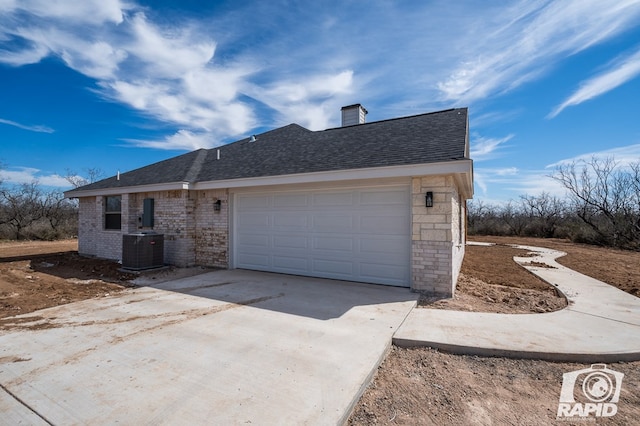 ranch-style home with an attached garage, central air condition unit, a shingled roof, concrete driveway, and a chimney