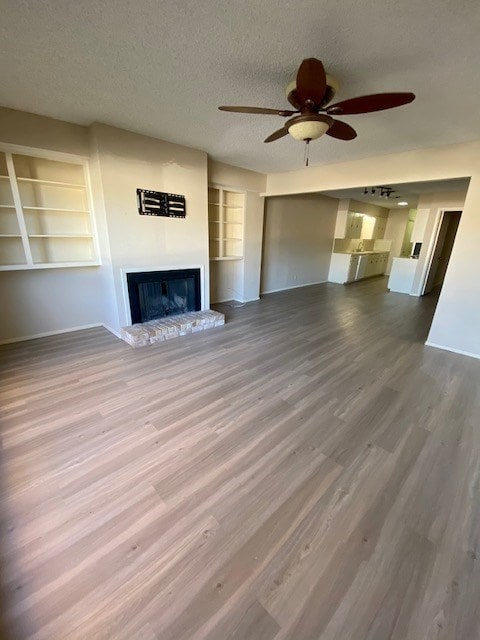 unfurnished living room with a textured ceiling, wood finished floors, a ceiling fan, built in features, and a glass covered fireplace