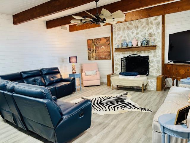 living room with wood finished floors, beamed ceiling, a stone fireplace, ceiling fan, and wooden walls