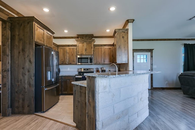 kitchen with appliances with stainless steel finishes, a kitchen island with sink, light stone counters, crown molding, and light hardwood / wood-style flooring