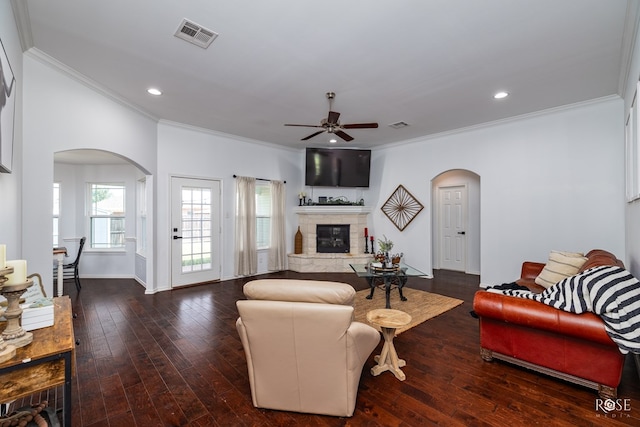 living room with crown molding, a stone fireplace, dark wood-type flooring, and ceiling fan