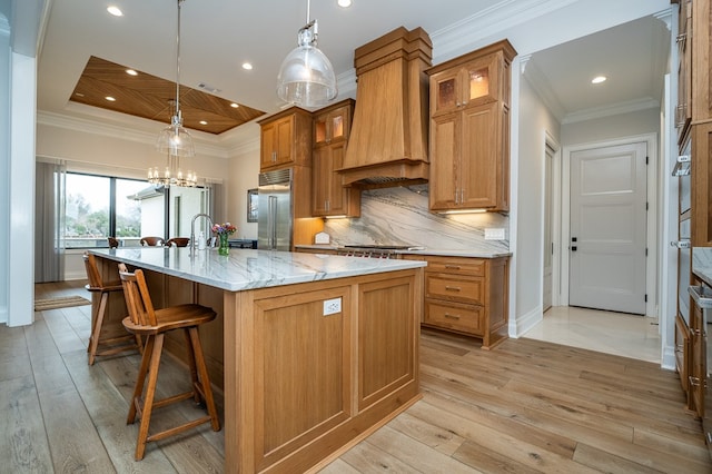 kitchen featuring appliances with stainless steel finishes, ornamental molding, custom range hood, an island with sink, and decorative light fixtures