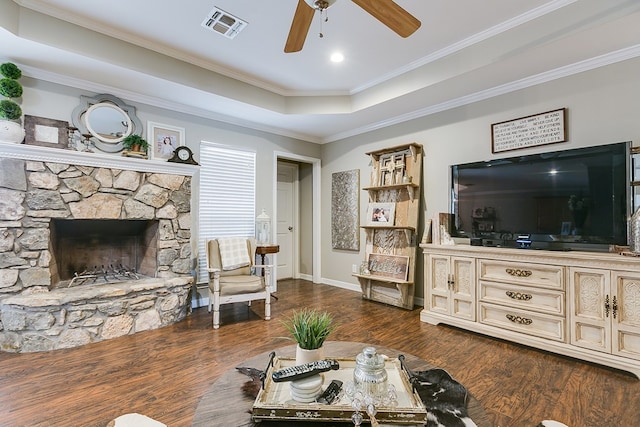 living room featuring dark wood-type flooring, ceiling fan, ornamental molding, a stone fireplace, and a raised ceiling