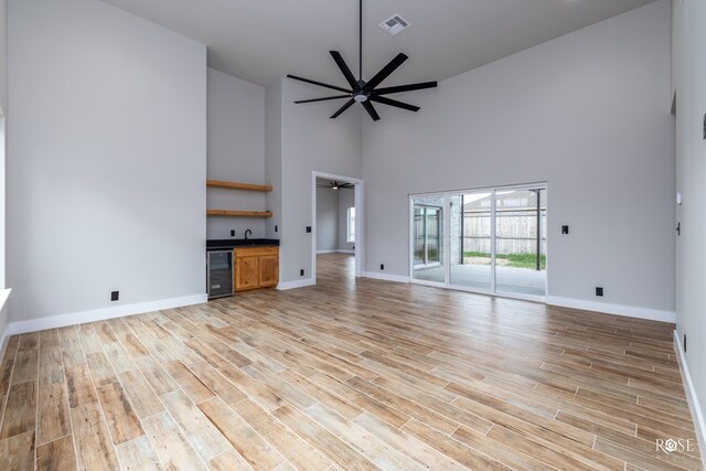 unfurnished living room featuring wine cooler, ceiling fan, light hardwood / wood-style floors, and a high ceiling