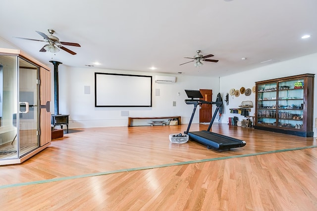 workout area featuring light wood-type flooring, a wall unit AC, ceiling fan, and a wood stove