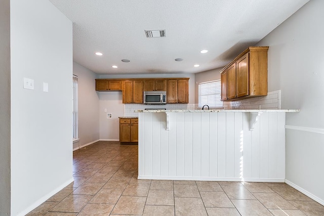 3622 Dominion Rdg, San Angelo, TX 76904 - kitchen with stainless steel microwave, brown cabinetry, tasteful backsplash, a peninsula, and a kitchen breakfast bar