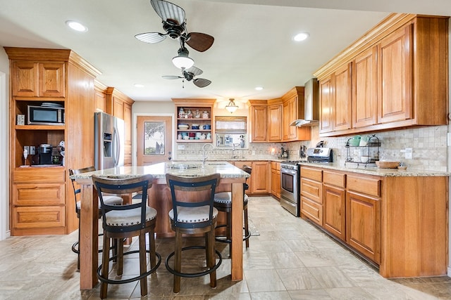 kitchen featuring a breakfast bar area, a kitchen island with sink, light stone counters, stainless steel appliances, and wall chimney range hood