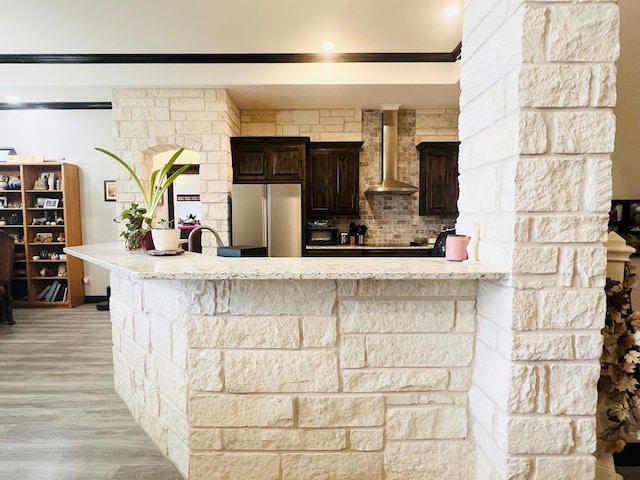 kitchen with light hardwood / wood-style flooring, stainless steel fridge, dark brown cabinets, kitchen peninsula, and wall chimney exhaust hood