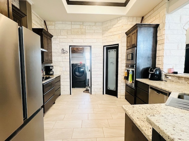 kitchen featuring dark brown cabinetry, a tray ceiling, stainless steel appliances, washer / clothes dryer, and light stone countertops