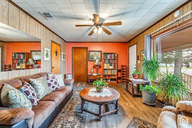 living area featuring wood walls, wood finished floors, visible vents, a ceiling fan, and crown molding