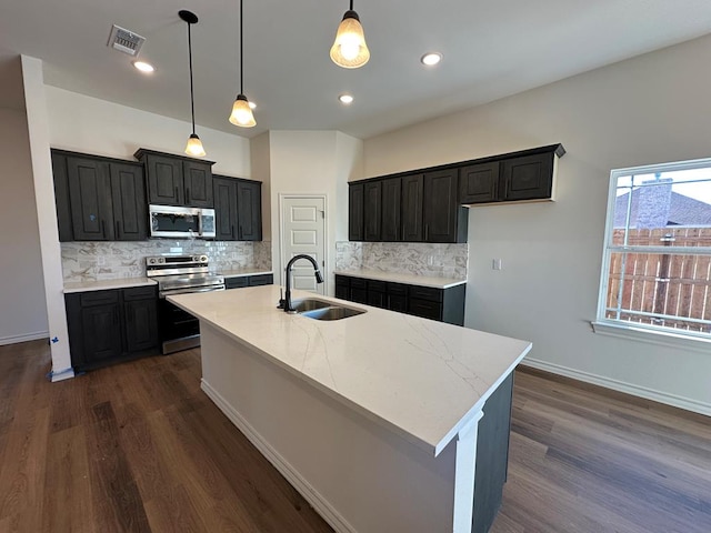 kitchen with decorative light fixtures, sink, stainless steel appliances, dark wood-type flooring, and a center island with sink