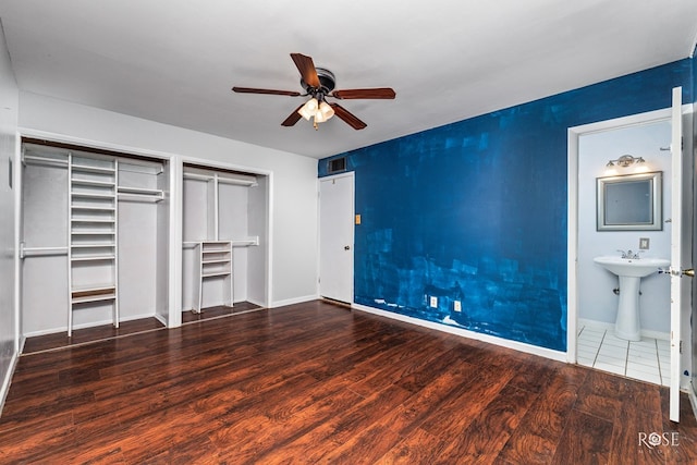 unfurnished bedroom featuring ensuite bathroom, sink, dark hardwood / wood-style flooring, two closets, and ceiling fan