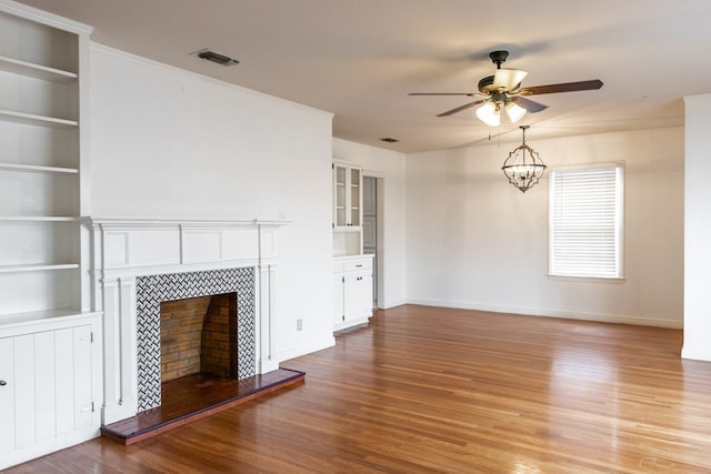 unfurnished living room with hardwood / wood-style flooring, built in shelves, ceiling fan with notable chandelier, and a tiled fireplace