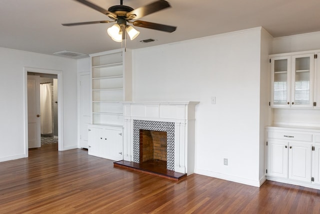 unfurnished living room featuring a tiled fireplace, built in shelves, dark hardwood / wood-style flooring, ceiling fan, and crown molding