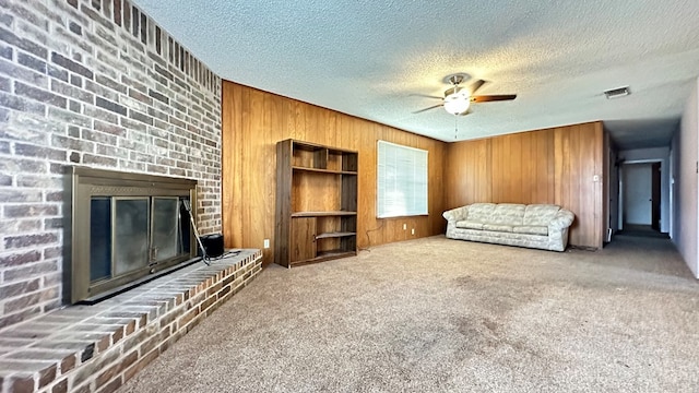 unfurnished living room with wooden walls, visible vents, ceiling fan, a brick fireplace, and carpet flooring