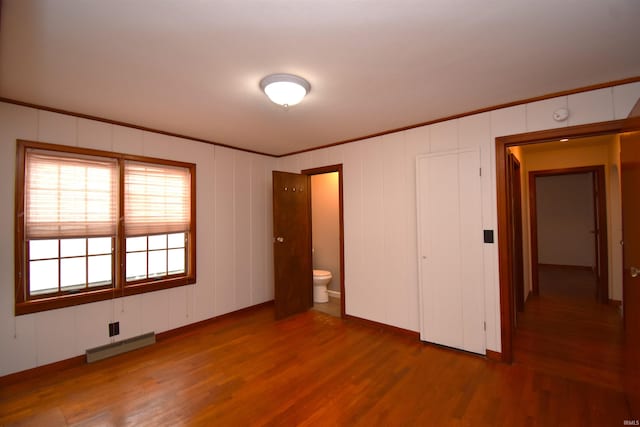 unfurnished bedroom featuring ornamental molding, ensuite bath, baseboard heating, and hardwood / wood-style flooring