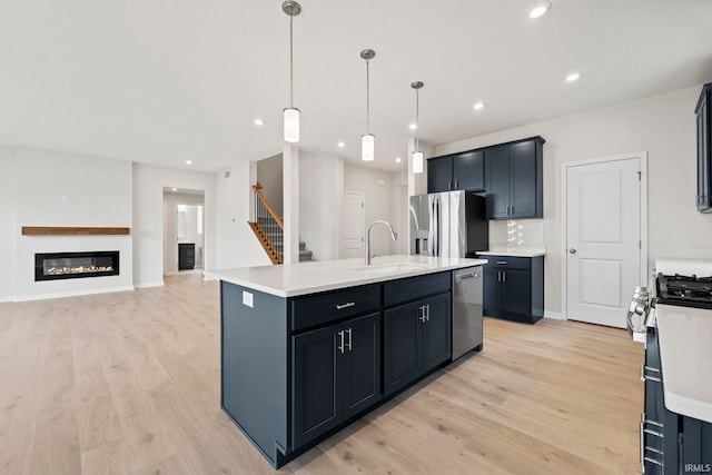 kitchen featuring an island with sink, appliances with stainless steel finishes, hanging light fixtures, light wood-type flooring, and sink