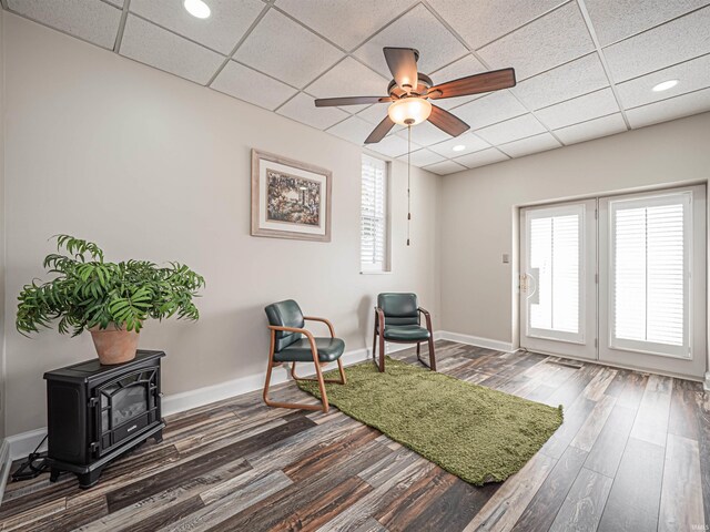 living area with a drop ceiling, a wood stove, ceiling fan, and dark hardwood / wood-style floors