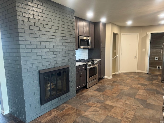 kitchen featuring light stone countertops, appliances with stainless steel finishes, a large fireplace, dark tile flooring, and dark brown cabinetry