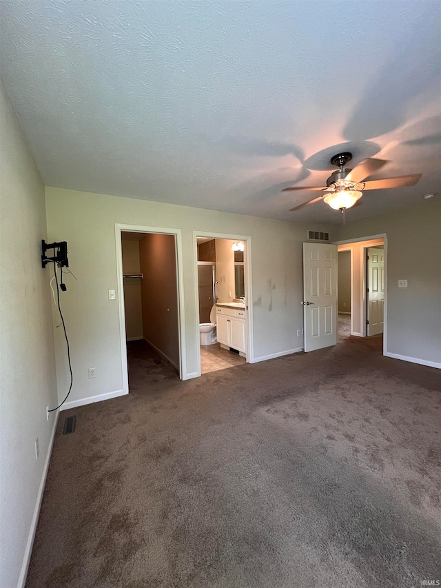 unfurnished bedroom with ceiling fan, a textured ceiling, connected bathroom, dark carpet, and a spacious closet