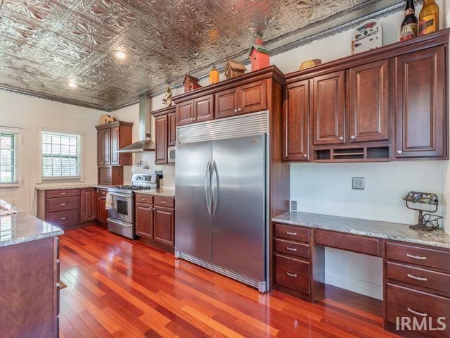 kitchen featuring wall chimney range hood, dark hardwood / wood-style floors, built in desk, light stone countertops, and appliances with stainless steel finishes