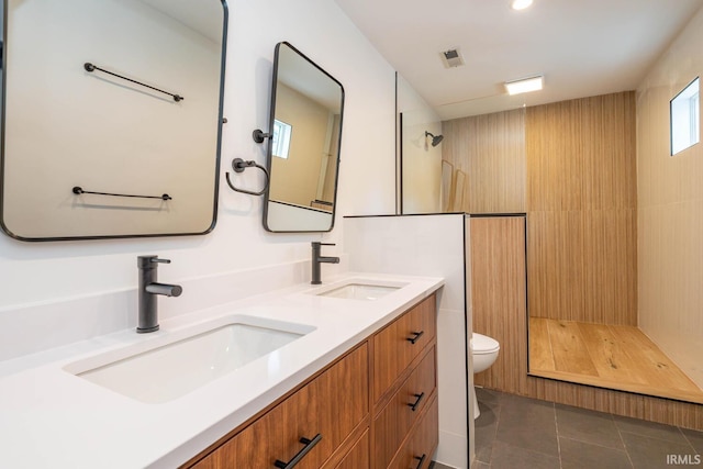 bathroom featuring tile flooring, tiled shower, double sink, vanity with extensive cabinet space, and toilet