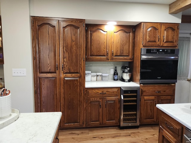 kitchen featuring decorative backsplash, light wood-style flooring, wine cooler, light countertops, and black oven